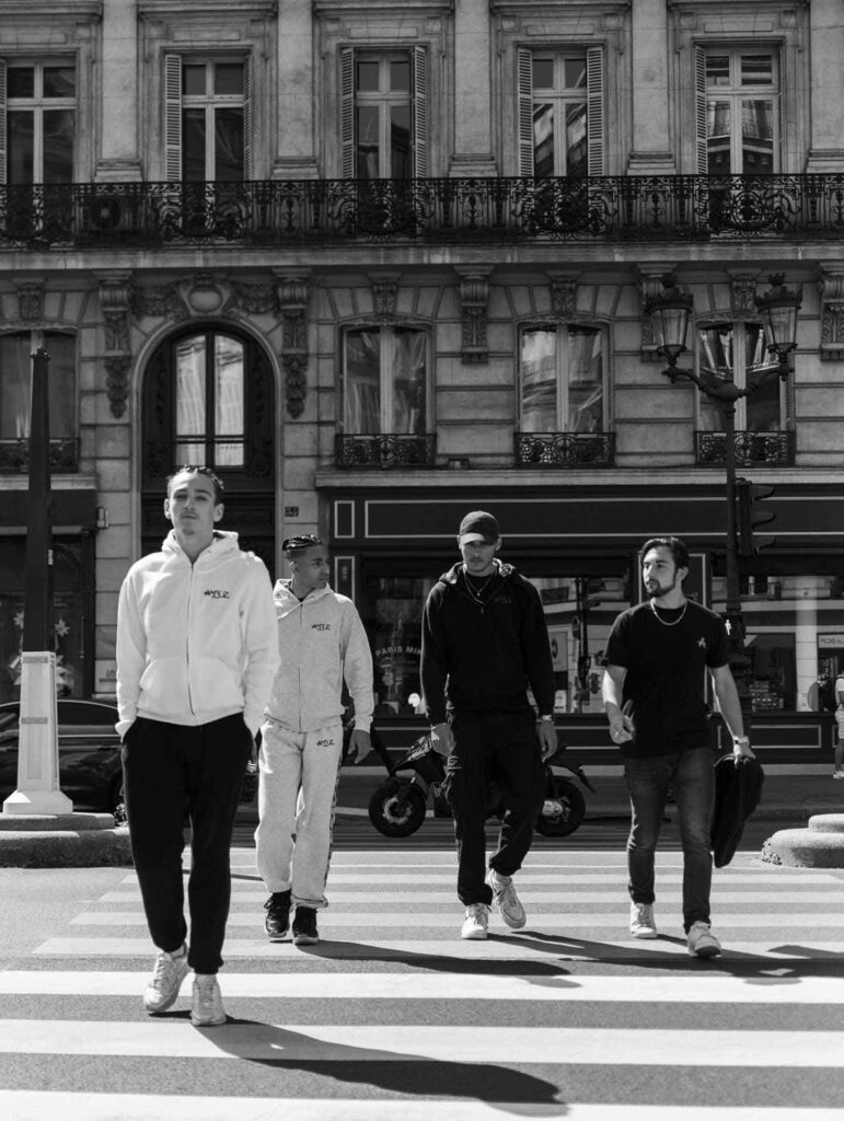 Image de la Collection Just Us d'Amble : un groupe d'amis marchant ensemble sur la route. Cette photo emblématique capture l'esprit de camaraderie et de style de vie urbain de la collection Just Us, désormais archivée.